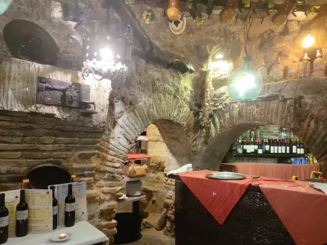 מרתף אבן מקומר עם בקבוקי יין, מנורות תלויות ושולחן במפה אדומה