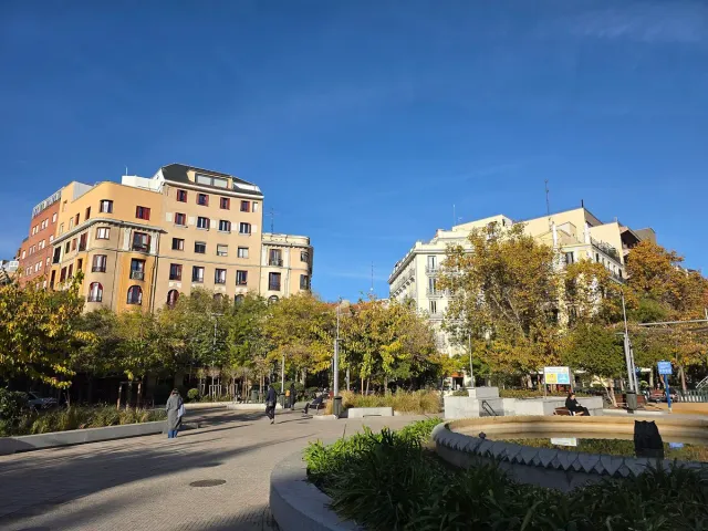 כיכר עגולה עם מזרקת אבן, ספסלים ועצי סתיו מול בתי מגורים בהירים