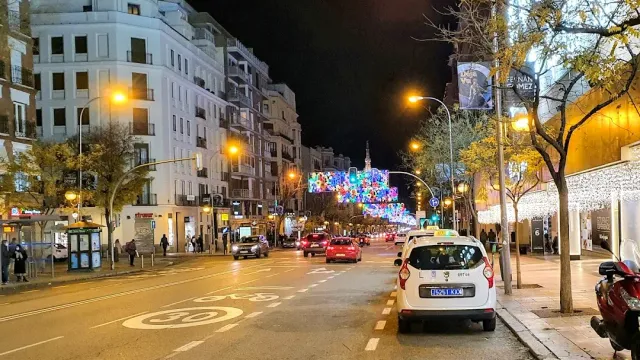 שדרה בלילה עם מכוניות, מונית חונה וקישוטי אור צבעוניים מעל הכביש