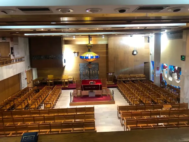 אולם עם שורות ספסלי עץ, במה מוגבהת בשטיח אדום ומנורת ויטראז׳ תלויה