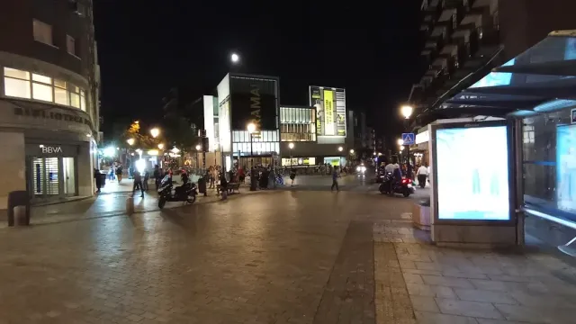 רחבה מרוצפת בלילה עם עוברי אורח, קטנועים חונים ושלטים מוארים