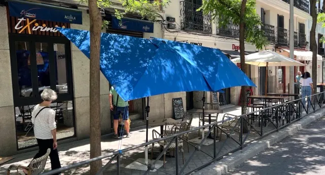 טרסת מדרכה עם שמשייה כחולה ושולחנות מתכת, ועוברי אורח ברחוב מוצל