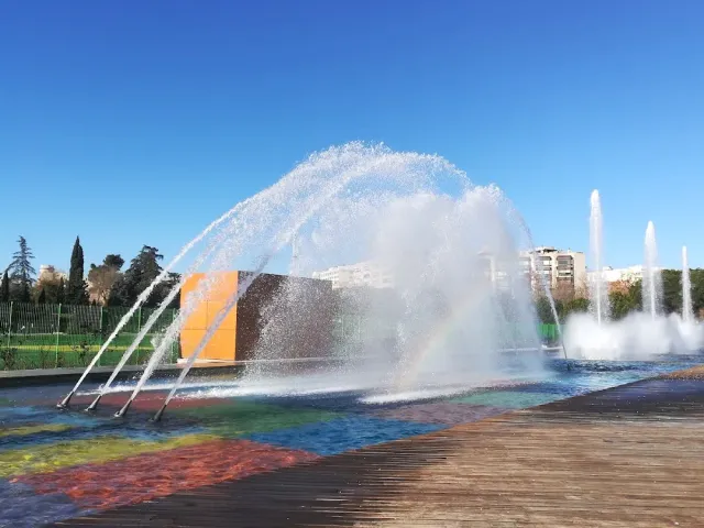 סילוני מים גדולים במזרקה עם קשת בענן בתרסיס, ורחבת עץ בחזית